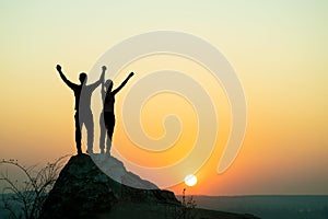 Man and woman hikers standing on a big stone at sunset in mountains. Couple raising up hands on high rock in evening nature.