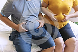 Man and woman holding their big belly while sitting on the bed suffering from extra weight. Couple heavy body size worry