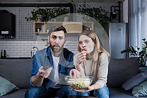 A man and a woman at home watching a boring movie, switching channels and eating popcorn