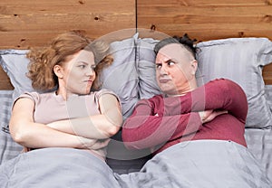 Man and a woman are lying in bed under a gray blanket, and they look at each other with displeasure.