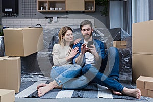 A man and a woman in a new apartment, a married couple sitting on the floor, near cardboard boxes, using the phone for online