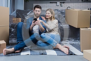 A man and a woman in a new apartment, a married couple sitting on the floor, near cardboard boxes, using the phone for online