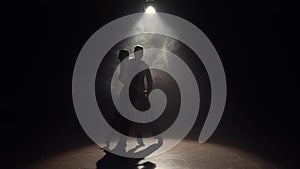 Man and woman perform Latin dance in darkness with overhead light and smoke. Silhouettes of pair of dancers passionately