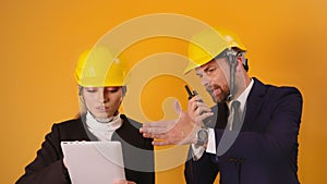 Man and woman professional architects with hard hats, tablet and walkie talkie isolated on orange background