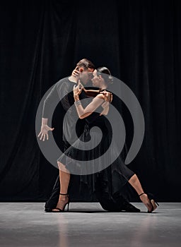 Man and woman, professional tango dancers performing in black stage costumes over black background. Attraction of