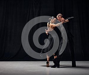 Man and woman, professional tango dancers performing in black stage costumes over black background. Elegance, romance