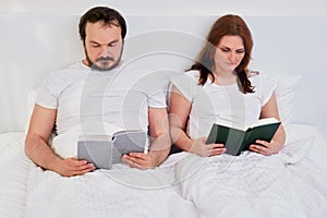 A man and a woman read books lying in a white bed, lifestyle. Husband and wife at home under coronavirus quarantine