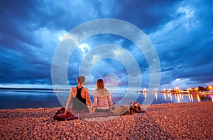 Man and woman resting on pebble beach at dusk on calm water dramatic blue cloudy sky background