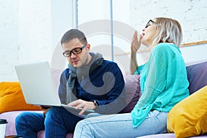 Man and woman sit on the couch, chatting and using a desktop computer.