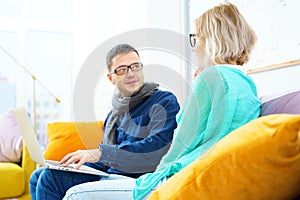 Man and woman sit on the couch, chatting and using a desktop computer.