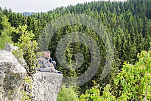 A man and a woman sit hugging each other on the edge of a mountain in nature