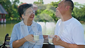 A man and a woman are sitting on a bench by a lake. The man is holding a cup of milk and the woman is holding a cup of coffee.