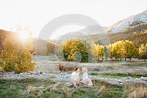 Man and woman are sitting on a blanket on a lawn in the middle of an autumn forest. Horse grazing in the meadow