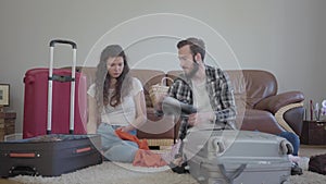 The man and woman sitting on the floor at home in front of leather sofa, packing a suitcase before travel. The husband