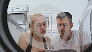 A man and a woman sitting in the house in front of a non-working washing machine studying the user manual.