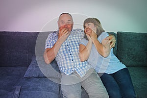 Man and a woman are sitting next to each other on a couch, enthusiastically watching a melodrama on television and empathizing, a