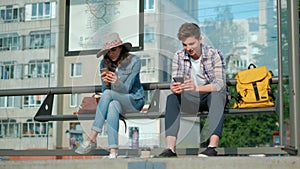 Man and woman are sitting together at transport station. They are holding modern gadgets and entertaining with internet