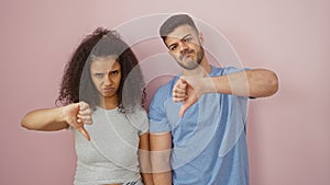 A man and woman stand against a pink background, both showing thumbs down with discontent expressions photo