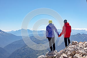 Man and woman stand holding hands on the edge of a cliff overlooking the mountains