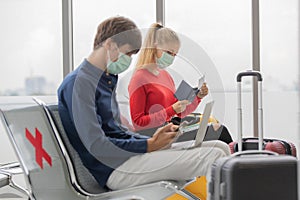 Man and woman travelers wearing protective hygiene masks sitting row seats with social distancing sign in airport lobby. They are