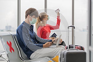 Man and woman travelers wearing protective hygiene masks sitting row seats with social distancing sign in airport lobby. They are