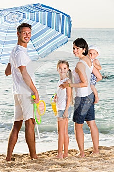 man and woman with two kids standing together under beach umbrella on beach