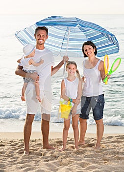 man and woman with two kids standing together under beach umbrella on beach