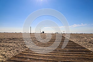 A man and a woman walking down a wooden boardwalk in the sand at the beach with people relaxing on the beach