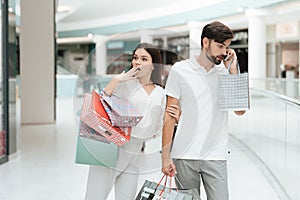 Man and woman are walking in shopping mall. Woman wants to go to store but man is busy talking on phone.