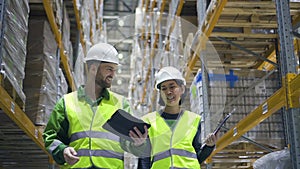 man and woman warehouse worker walk through storage racks with merchandise and look at digital tablet