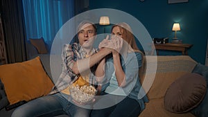 A man and a woman are watching a horror movie while sitting on the couch with a plate of popcorn. A woman in fear