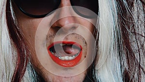 Man in woman wig in sunglasses say words in camera. Red pomade on lips. Parody