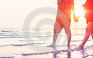 The man and the women hold one`s hand on the beach and front of sunset. the women wear red dress. thay are couple. they are walkin