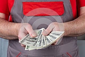 A man in work clothes holds a stack of 100 dollar bills in his hands