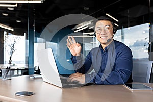 Man at work with headset phone and laptop successful asian man smiling and looking at camera at workplace inside office
