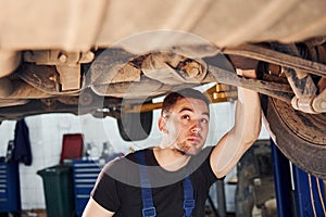 Man in work uniform standing under car and repairs it indoors. Conception of automobile service