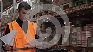 man worker employee in orange vest walking along warehouse checking papers verification of with boxes of products with