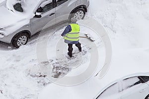 Man, worker in green safety vest shoveling snow near cars on road, parking