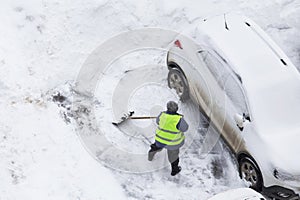 Man, worker in green safety vest shoveling snow near cars on road, parking