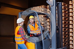 Man worker sitting in industrial forklift at warehouse talking to woman co-worker