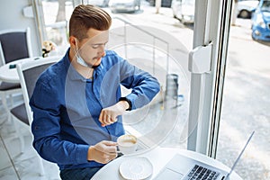 Man working from a cafe checking time on his watch and drinking coffee. Businessman wearing sterile medical mask leading matters