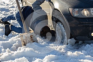 Man working at car stuck in snow on knee with shovel at daylight offroad