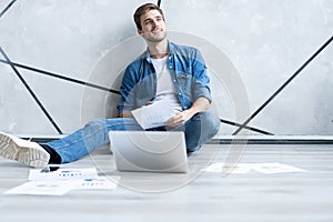 Man working at home. Handsome young man sitting on the floor and examining document while laptop and documents laying