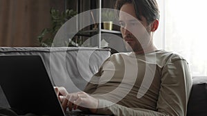 A man is working from home lying on the couch, thinking and typing on his laptop