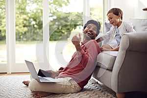 Man Working From Home Sitting On Floor In Lounge Using Laptop And Mobile Phone As Son Watches