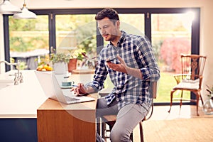 Man Working From Home Using Laptop On Kitchen Counter Talking Into Microphone On Mobile Phone