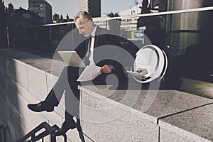 A man is working on a laptop, sitting on a parapet. Near his mono-wheel