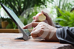 Man working on tablet computer with bitten apple in his hand. Wooden table. Green garden background.