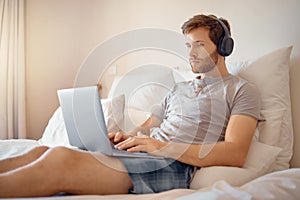 Man working or typing on laptop and listening to music with headphone in bed at home. Remote worker streaming podcast