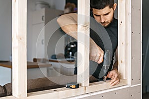 The man works as a screwdriver, fixing a wooden frame for the window to the gypsum plasterboard partition.